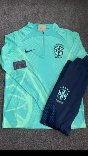 Nike Brasil långärmad träningströja - Storlek - 150-165. Snygg långärmad träningströja från Nike med Brasil-logga på bröstet. Turkosblå färg med ljusa grafiska mönster och halv dragkedja vid halsen. Tillverkad i lätt och ventilerande funktionsmaterial, perfekt för fotbollsträning.