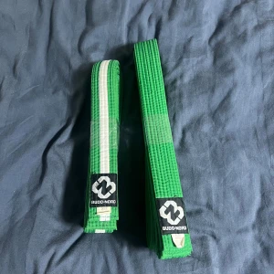 Gröna Budo-Nord bälten för kampsport - Två gröna bälten från Budo-Nord, ett med vit rand och ett helgrönt. Perfekta för kampsport som judo eller karate. Bältena är tillverkade i en blandning av bomull och polyester och har klassisk design.