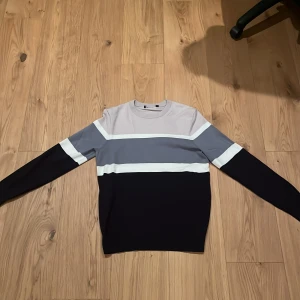 Randig långärmad tröja i blått och grått - Snygg långärmad tröja med breda horisontella ränder i ljusgrått, blått, vitt och mörkblått. Tröjan har en klassisk rund halsringning och är perfekt för dig som gillar stilrena färgblock. Passar till jeans eller chinos för en clean look.