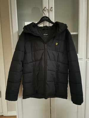 Svart pufferjacka från Lyle & Scott - Svart pufferjacka från Lyle & Scott med huva och gul logga på bröstet. Jackan har quiltad design, två fickor med dragkedja och hel dragkedja framtill. Perfekt för kalla dagar och har en clean, stilren look. Jackan har två små hål men går enkelt att laga med små laggninglappar vilket inte kommer påverka utseendet.