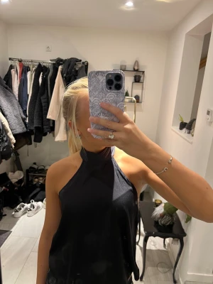 Svart halterneck topp med hög krage - Säljer en svart halterneck topp med hög krage och ärmlös design. Toppen har en stilren och enkel look med lätt glansigt tyg som faller snyggt. Perfekt för dig som gillar minimalistisk stil och vill ha något som sticker ut med sin höga halsringning.