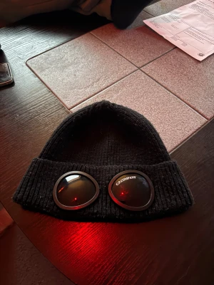 Svart C.P. Company Goggle Beanie - Säljer en svart ribbstickad mössa från C.P. Company med ikoniska goggle-linser framtill. Mössan är tillverkad i 100% bomull och har en bred uppvikt kant. Perfekt för dig som vill sticka ut med streetwear-vibe.