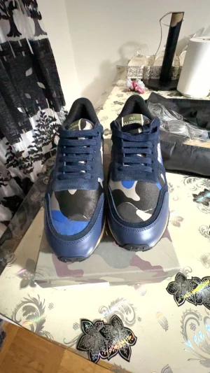 Valentino Rockrunner - Säljer ett par Valentino Rockrunner sneakers med blå, svart och beige camouflage-mönster. Skorna har snörning, platt sula och rund tå. Materialmix med skinn och textil, samt detaljer i mocka. Perfekt för dig som gillar statement-sneakers med lyxig vibe.