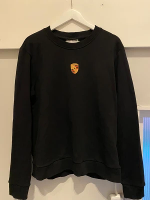 Svart sweatshirt från Porsche - Svart sweatshirt från Porsche som aldrig har använts. Storlek L med klassiskt rund hals och ribbade muddar. På bröstet sitter ett snyggt Porsche-emblem i gult, rött och svart. Tröjan är tillverkad i mjuk bomull och har lång ärm för en chill och stilren look.  (Pris kan diskuteras vid smidig affär)