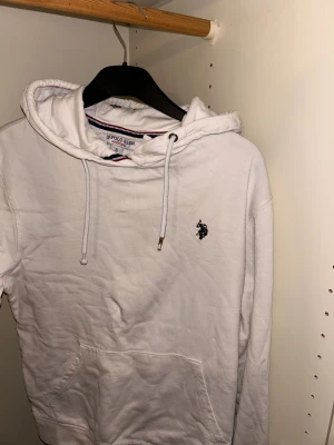 Vit hoodie från U.S. Polo Assn. - Vit hoodie från U.S. Polo Assn. med klassisk broderad logga på bröstet. Tröjan har huva med snörning, känguruficka och ribbade muddar. Mjuk bomullskvalitet och stilren look, perfekt för chill dagar. Snygga detaljer med metalländar på snöret.