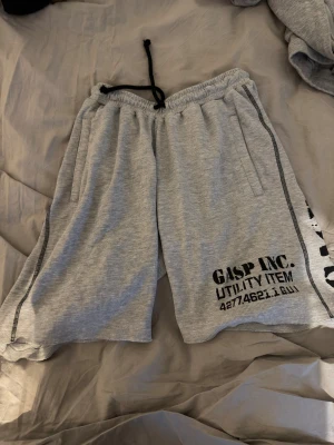 Grå mesh shorts från GASP INC. med tryck - Säljer ett par grå mesh shorts från GASP INC. med svart tryck på benet och utility-inspirerad text. De har elastisk midja med svart snörning och två sidofickor. Materialet är mjukt och ribbat, perfekt för chill eller träning. Nypris 799kr
