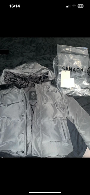 Grå dunjacka Canada Goose  - Säljer en grå dunjacka från Canada Goose, Jackan har en klassisk puffer-design, svarta detaljer, logotyp på ärmen och praktiska fickor. Perfekt för kalla vinterdagar och har både dragkedja och knappar framtill.