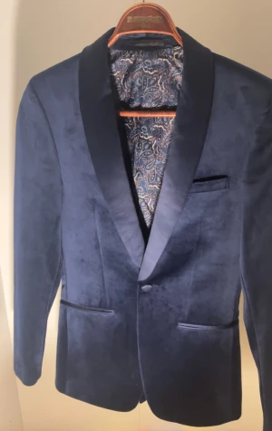 Mörkblå sammetskavaj med paisleyfoder - Snygg mörkblå kavaj i sammet med breda slag och paisleymönstrat innerfoder. Klassisk passform med två fickor fram och en bröstficka. Perfekt för dig som vill sticka ut med en elegant och lyxig look.