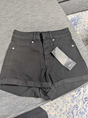 Svarta shorts - Svarta shorts från FB Sister i storlek XS, med klassisk femficksdesign och uppvikta benslut. Med bälteshällor och silverfärgade nitar. Snygg, enkel stil som funkar till många looks. Materialet är mjukt och stretchigt, troligen bomull med inslag av syntet. Nypris 199