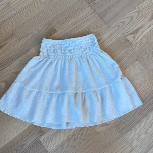 Vit volangkjol från Gina Tricot - Supersöt vit kortkjol från Gina Tricot i storlek XXS. Kjolen har smockad midja och två lager med volanger som ger en flowy och chill vibe. Materialet är mjukt och lätt, perfekt för varma dagar. Enkel att matcha med crop tops eller hoodies för en fräsch look.