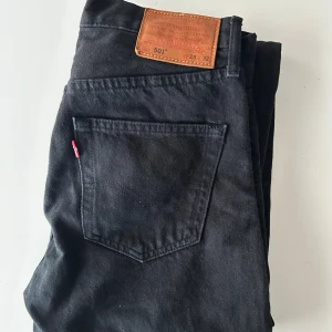Svarta Levi's 501 jeans W28 L32 - Svarta Levi's 501 jeans med klassisk femficksdesign och rakt snitt. Jeansen har en brun läderpatch med logga bak i midjan och den ikoniska röda Levi's-taggen på bakfickan. Tillverkade i slitstarkt bomullstyg, perfekta för en cool och tidlös look.