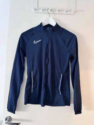 Marinblå Nike zip-tröja - Snygg marinblå långärmad zip-tröja från Nike med vit logga på bröstet och vita detaljer vid fickorna. Perfekt för träning eller chill, med smal passform och hög krage. Materialet är mjukt och känns sportigt.