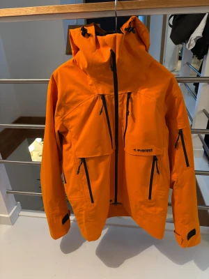 Skidjacka  - Snygg orange vindjacka från Everest med huva och flera svarta dragkedjor på bröstet och ärmarna. Jackan har justerbara muddar med kardborre och praktiska fickor framtill. Perfekt för dig som gillar att sticka ut och vill ha en jacka med coola detaljer.