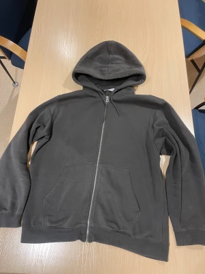Svart zip-hoodie från Weekday - En svart hoodie från Weekday med dragkedja hela vägen, huva med dragsko och två stora fickor framtill. Tillverkad i mjuk bomullsblandning och har ribbade muddar vid ärmslut och nederkant. Enkel och clean stil som funkar till allt.