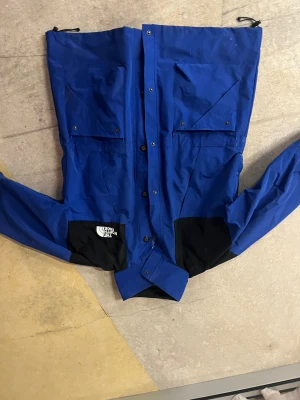 Blå vinterjacka från The North Face - SLÅ ETT BUD!!  Blå och svart vindjacka från The North Face i storlek L, tillverkad i slitstark GORE-TEX. Jackan har stora fickor med knappar, hög krage och logga på både fram- och baksida. Perfekt för dig som gillar outdoor och streetstyle.