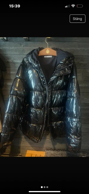 Svart jacka från Zara - Säljer en svart, glansig pufferjacka från Zara i storlek L. Jackan har huva, dragkedja och breda, quiltade sektioner som ger en cool och voluminös look. Perfekt för kalla dagar när du vill ha både stil och värme.