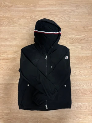 Svart vindjacka från Moncler - Svart vindjacka från Moncler med huva och snygg rödvit rand på kanten. Jackan har dragkedja framtill, två fickor med silverknappar och elastiska muddar vid ärmslut och nederkant. Moncler-logga på vänster ärm. Perfekt för blåsiga dagar.