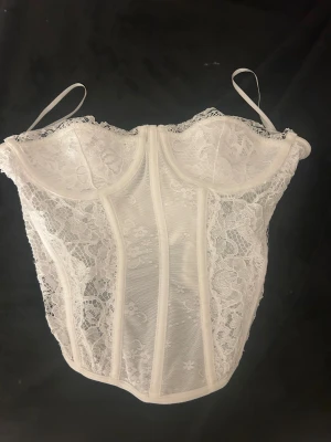 Vit spetskorsett med axelband - Säljer en vit korsett med vacker spets över hela toppen. Den har markerade sömmar och blommigt spetsmönster som ger en romantisk vibe. Perfekt för dig som vill ha en feminin och trendig look.