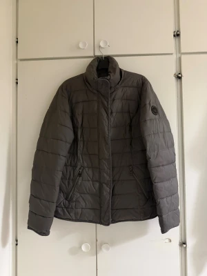 Grå pufferjacka från Street One - Snygg grå pufferjacka från Street One med hög krage och två praktiska fickor med dragkedja. Jackan har quiltad design och ett svart märke på ärmen. Perfekt för kyliga dagar och har en stilren look som funkar till många outfits.