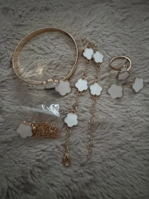 Smyckesset med vita blommor i guld - Säljer ett komplett smyckesset med armband, halsband, örhängen, ring och armlänk i guldfärgad metall. Alla delar har vita blommor i pärlemoliknande material som ger en lyxig och trendig känsla. Perfekt för dig som gillar stilrena accessoarer med en söt twist.