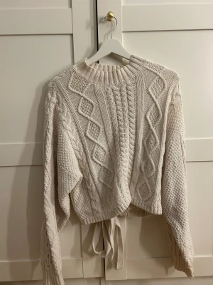 Beige stickad tröja med knyt - Säljer en beige stickad tröja med grova flätmönster och ribbad hals. Tröjan har långa ärmar och ett snyggt knyt i midjan som ger en chill vibe. Perfekt för dig som gillar chunky stickat och vill ha något mysigt och trendigt. (har två små fläckar på baksidan)