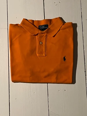Orange pikétröja från Polo Ralph Lauren - Snygg orange pikétröja från Polo Ralph Lauren med klassisk krage och två knappar framtill. Tröjan har det ikoniska blå broderade Polo-logot på bröstet och är tillverkad i 100% bomull för skön komfort. Perfekt för dig som gillar färg och stil.