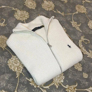 Vit stickad kofta från Polo Ralph Lauren - Vit ribbstickad kofta från Polo Ralph Lauren med hel dragkedja och klassisk krage. Tröjan har broderad marinblå logga på bröstet och är tillverkad i ett stickat material. Perfekt för lager-på-lager och stilren look.