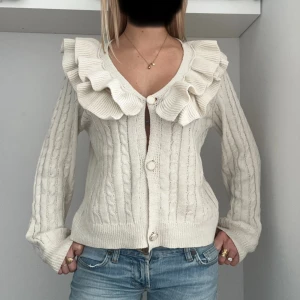 Beige kofta med volangkrage - Superfin beige kofta med grovstickat kabelmönster och stora volanger runt v-ringad hals. Koftan har knappar framtill och långa ärmar. 💕 det är ett litet hål på sidan av tröjan skriv privat för bild på detta 