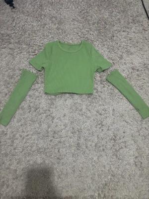 Grön ribbad croppad topp - Säljer en ljusgrön croppad topp med ribbad struktur och rund hals. Toppen har korta ärmar och kommer med matchande avtagbara ärmar för en extra cool look. Perfekt för dig som gillar att mixa stilar och sticka ut.