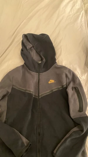 Nike hoodie med dragkedja och huva - Snygg svart och mörkgrå hoodie från Nike med gul logga på bröstet. Hoodien har hel dragkedja, huva och en praktisk ficka med dragkedja på ena ärmen. Perfekt för chill eller träning. Mjukt material som känns skönt mot huden.