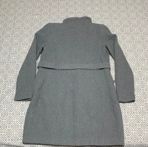 Grå kappa med bälte och dragkedjor - Snygg grå kappa med hög krage, bälte i midjan och svarta detaljer vid fickorna. Kappan har långa ärmar och stängs med både dragkedja och knappar. Perfekt för dig som gillar stilrena och moderna plagg med lite edge.