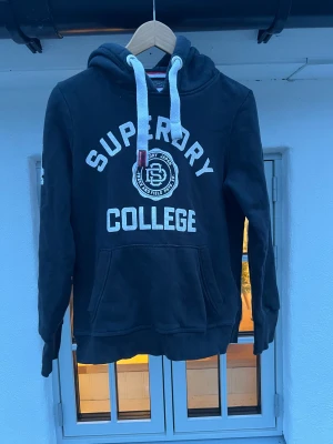 Svart Superdry College hoodie - Svart hoodie från Superdry med vit college-tryck på bröstet och siffran 54 på ärmen. Stor ficka framtill, vit dragsko och broderad logga på huvan. Mjuk bomullsmix, klassisk passform och sportig vibe. Perfekt för chill dagar och streetstyle.