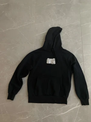 Svart hoodie från Mavi Black Pro - Svart hoodie från Mavi Black Pro i storlek S. Tröjan har en stor huva med snörning, magficka och en vit patch med text på bröstet. Tillverkad i mjuk bomull och polyesterblandning, perfekt för chill dagar. Klassisk loose passform och långa ärmar.