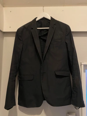 Svart kavaj/blazer från Acne Studios - Snygg svart kavaj/blazer från Acne Studios med klassisk passform och stilrena detaljer. Kavajen har två fickor med lock, bröstficka och innerficka med knapp. Fodret är ljust och materialet känns lätt och smidigt. Perfekt för dig som gillar minimalistisk design. En liten reva i fodret på ena armen men det ser man inte. (Pris går att diskutera vid smidig affär)