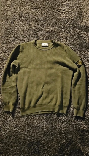 Grön stickad tröja från Stone Island - Snygg olivgrön stickad tröja från Stone Island med klassisk rund hals och ribbade muddar. Tröjan har lång ärm och den ikoniska Stone Island-loggan på vänster ärm. Perfekt för dig som gillar streetwear och vill ha en clean look.
