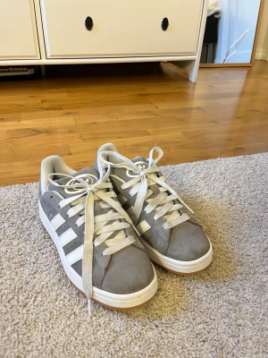 Grå Adidas campus 00 unisex - Säljer ett par grå adidas campus sneakers med klassiska vita ränder på sidorna och vit logga på plösen. Skorna har rund tå, platt sula och snörning. Ovandelen är i mocka och insidan är mjukt vadderad för extra komfort. Perfekt för dig som gillar stilrena och snygga skor. Skorna kommer även med ett par gråa snören. Knappt använda. Säljs på grund av att det var fel storlek för mig. 
