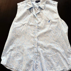 Blommig ärmlös skjorta från Polo - Snygg ljusblå ärmlös skjorta från Polo med blommigt mönster och klassisk krage. Skjortan har knappar framtill och ett broderat Polo-logotyp på bröstet. Perfekt för dig som gillar stilrena och fräscha plagg med lite extra detaljer.