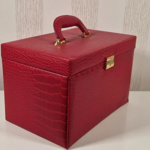 Röd smyckesväska i krokopräglat skinn - Snygg röd smyckesväska med krokopräglat mönster och guldfärgade detaljer. Väskan har flera fack, lådor och spegel på insidan, perfekt för att organisera smycken och accessoarer. Praktiskt handtag upptill gör den enkel att bära med sig.