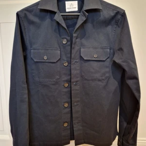 Mörkblå overshirt från The Shirt Factory - Snygg mörkblå overshirt från The Shirt Factory. Storlek 37 motsvarande S, nyskick. Skjortan har klassisk krage och två bröstfickor med knappar. Jackan har långa ärmar och är tillverkad i bomull. Perfekt att slänga på över en hoodie eller t-shirt. Nypris:1699