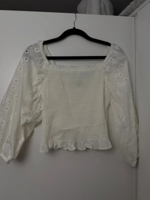 Vit blus med broderade ärmar - Supersöt vit blus med smockad kropp och volangkant nertill. Ärmarna är långa och puffiga med broderade blommor och små hål, vilket ger en romantisk vibe. Fyrkantig ringning och lätt struktur i tyget. Perfekt för vår och sommar!