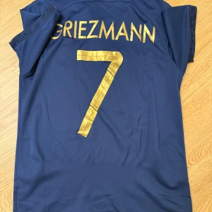 Frankrike Griezmann #7 Nike fotbollströja - Säljer en officiell fransk landslagströja från Nike med Griezmann och nummer 7 i guld på ryggen. Tröjan är marinblå med korta ärmar, guldiga detaljer och FFF-logga på bröstet. Materialet är lätt och andas, perfekt för match eller träning.