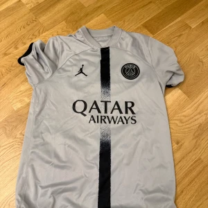 PSG Mbappé grå fotbollströja - Snygg grå PSG fotbollströja med svart vertikal rand på framsidan, Jordan-logga och Paris Saint-Germain-emblem. Tryck med Qatar Airways på bröstet och Mbappé samt nummer 7 på ryggen. Kortärmad modell i lätt och ventilerande material.