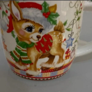 Mugg Ren Motiv  - Bilderna visar en julmugg med ett sött renmotiv, perfekt för varma drycker under vintern. Muggen har färgglada detaljer med en ren i tomteluva och halsduk, samt juldekorationer. Inga synliga skador eller slitage. Passar utmärkt som present eller till julfikat.