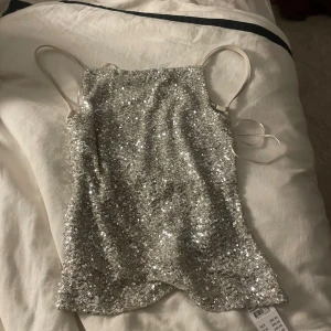 Glittrig silver paljett-topp från Ginatricot - Säljer en glittrig silverfärgad topp från Ginatricot med tunna axelband och croppad passform. Toppen är täckt av paljetter på framsidan och har ett mjukt, beige foder på insidan. Perfekt för att sticka ut med extra bling och snyggt skimmer.