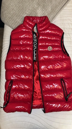 Moncler väst - Snygg röd dunväst från Moncler med glansig finish och svarta detaljer. Västen har två fickor med dragkedja, hög krage och ett broderat Moncler-märke på bröstet. Insidan har ett coolt serietidningsmotiv och tydlig Moncler-logga längs dragkedjan. Aldrig använt 