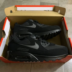 Nike Air Max 90 Svarta sneakers - Nike Air Max 90 sneakers i helsvart med grå swoosh och detaljer. Skorna har en robust platt sula med synlig Air Max-enhet, snörning och rund tå. Ovandelen är i syntet och textil, vilket ger en sportig och clean look. Perfekt för dig som gillar klassisk streetstyle.