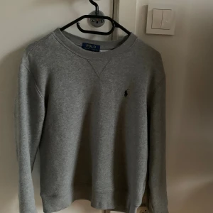 Grå sweatshirt från Polo Ralph Lauren - Snygg grå sweatshirt från Polo Ralph Lauren med klassisk rund hals och diskret logga broderad på bröstet. Tröjan har långärmade muddar och är gjord i mjuk bomullsmix som är perfekt för chill dagar. Enkel och stilren design som funkar till jeans eller joggers.