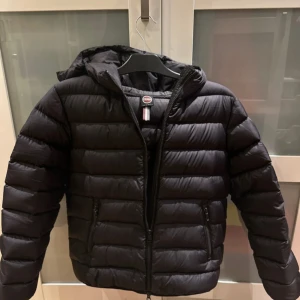 Svart Colmar pufferjacka med huva - Säljer en svart pufferjacka från Colmar Originals med huva och dragkedja framtill. Jackan har horisontella quiltade sektioner och två sidofickor med dragkedja. Perfekt för kalla dagar och har en clean, sportig look.