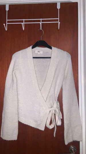 Vit omlottkofta från Gina Tricot XS - Supermysig vit omlottkofta från Gina Tricot i ullblandning. Koftan har långa ärmar, ribbade muddar och knytband i midjan för en snygg passform. Perfekt för lager-på-lager och chilliga dagar. Enkel och stilren design som är lätt att matcha.