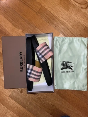 Burberry sandaler rutig svart/beige - Snygga sandaler från Burberry med klassiskt rutigt mönster i beige, svart, vitt och rött på remmen. Svart platt sula i syntetmaterial, med Burberry-logga på undersidan. Levereras med originalkartong och dustbag. Perfekt för sommaren!